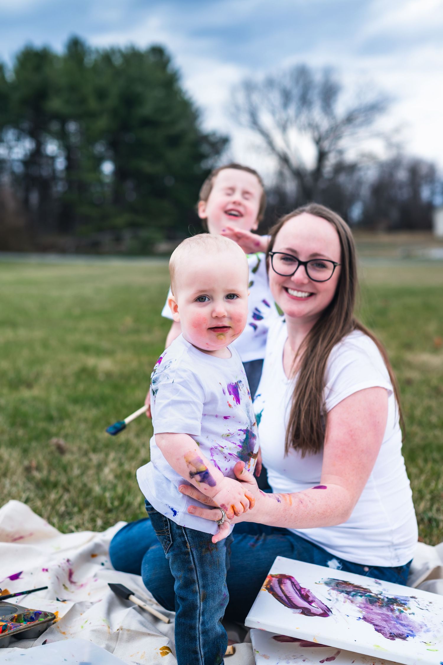 Fun outdoor painting activity captured with paint-splashed clothes and happy expressions against an evergreen backdrop.
