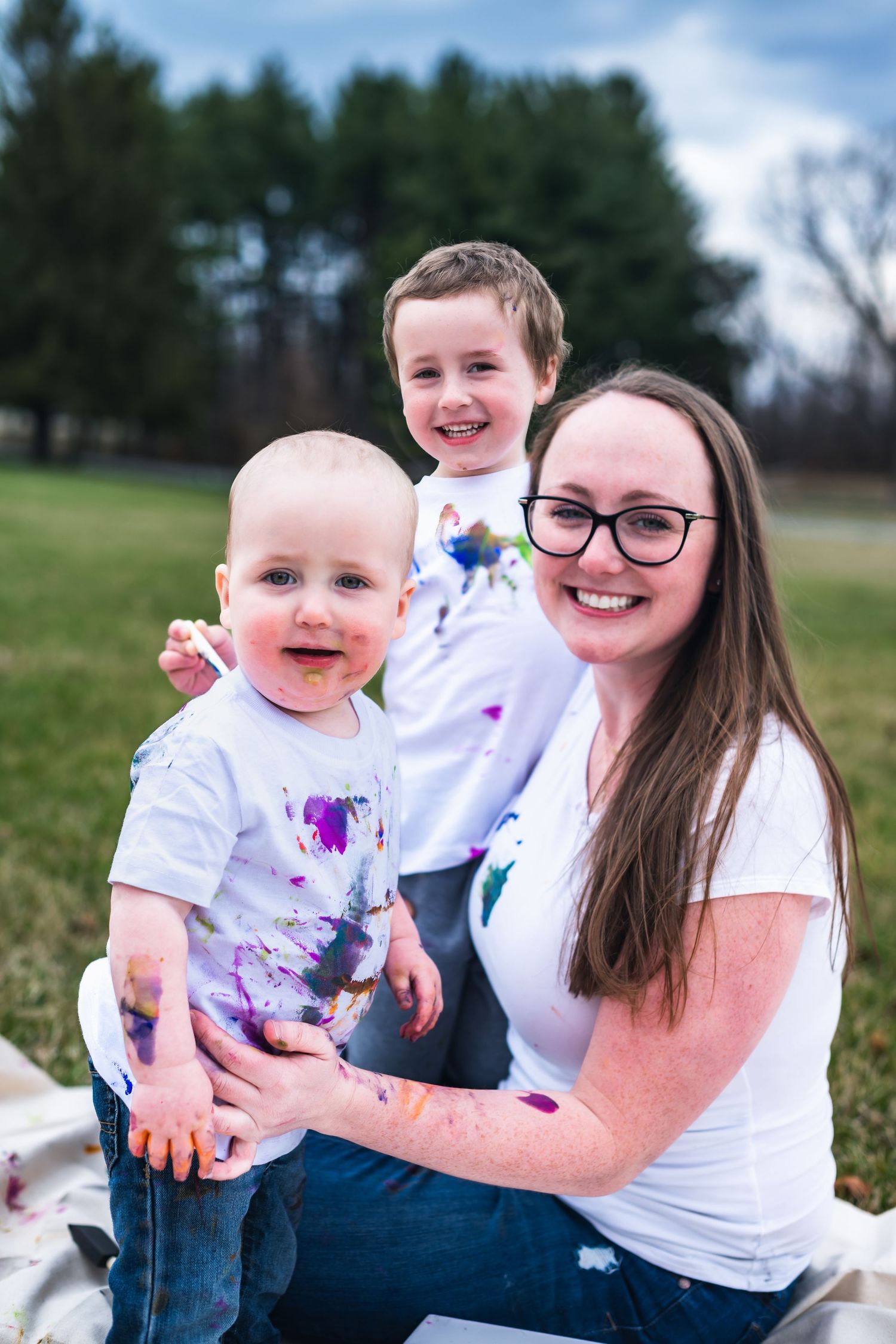 A joyful outdoor family photo featuring playful paint-splattered white shirts and smiles on a sunny day in a grassy field.