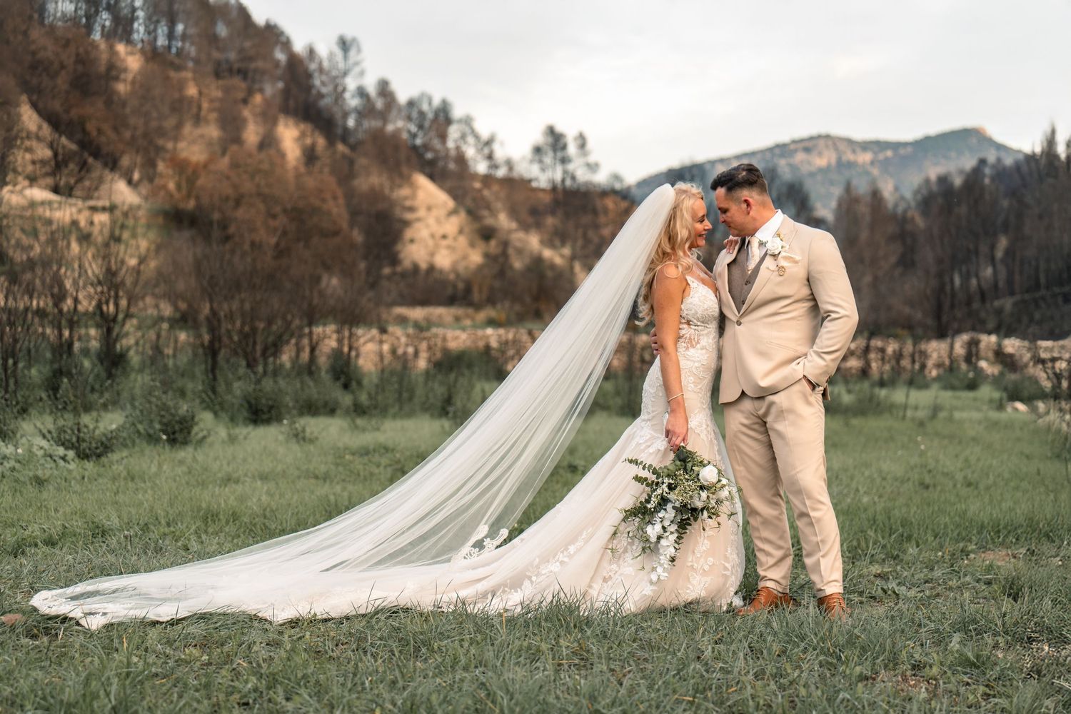 Couple looking at each other in wedding attire wedding dress with long veil in Spanish country side with mountains in the background