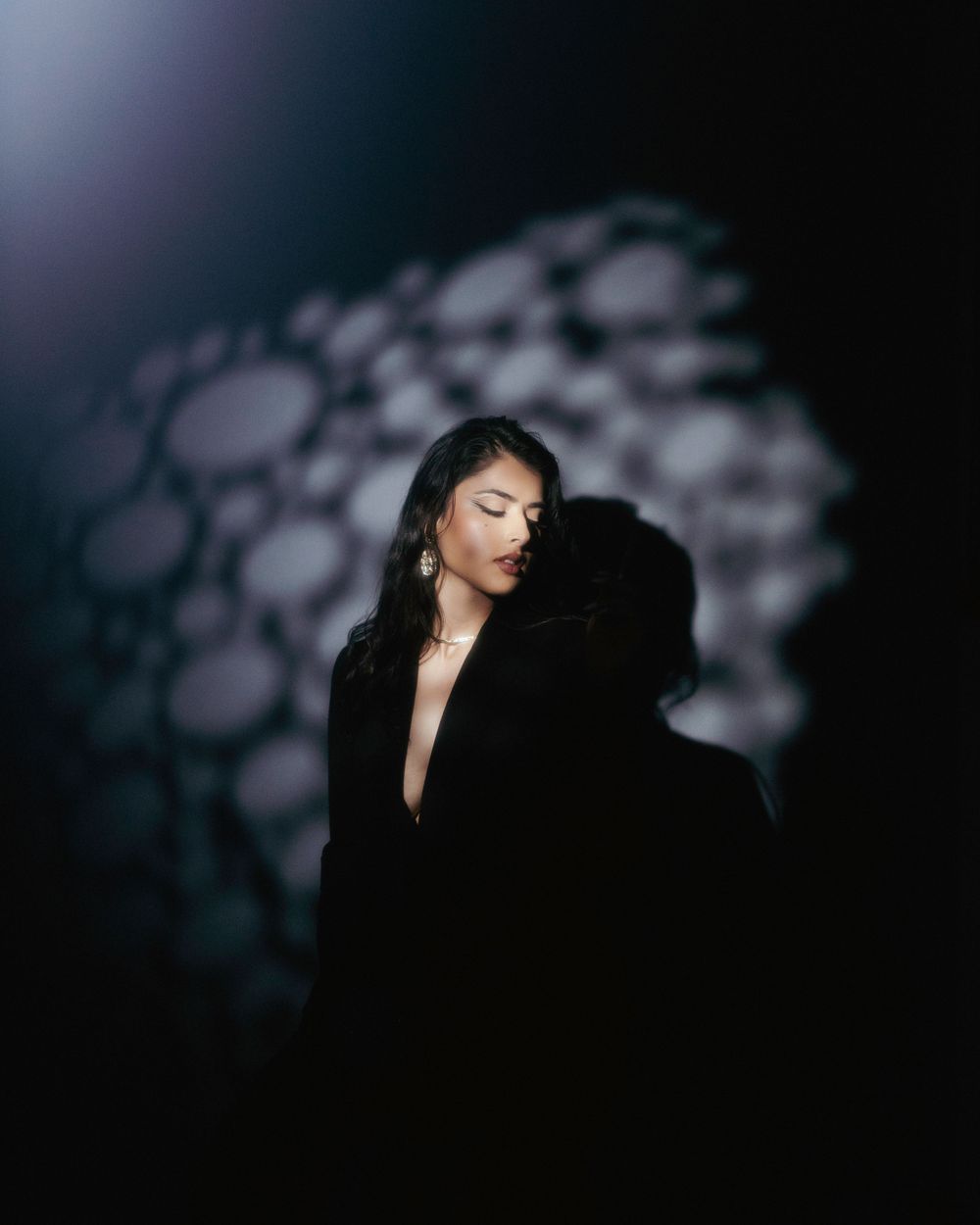 Figure in a black plunging neckline dress poses dramatically against blurred bokeh lights in dark setting.