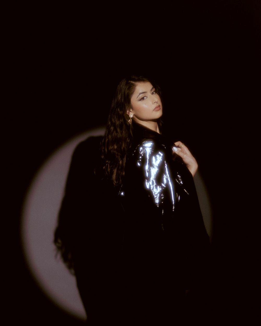 Dramatic monochrome portrait with a shimmering silver jacket against dark backdrop with crescent moon shadow.