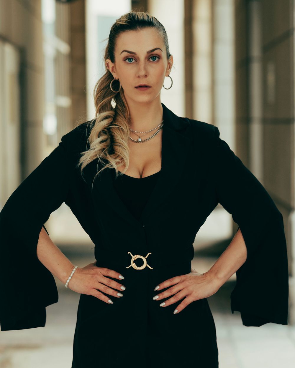 Professional fashion photo showing a confident pose in a sleek black dress with dramatic bell sleeves and statement jewelry.