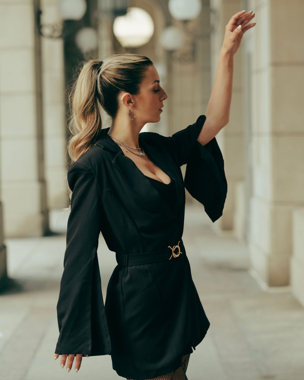 Elegant figure in a black belted blazer dress with bell sleeves strikes a graceful pose in a columned hallway.