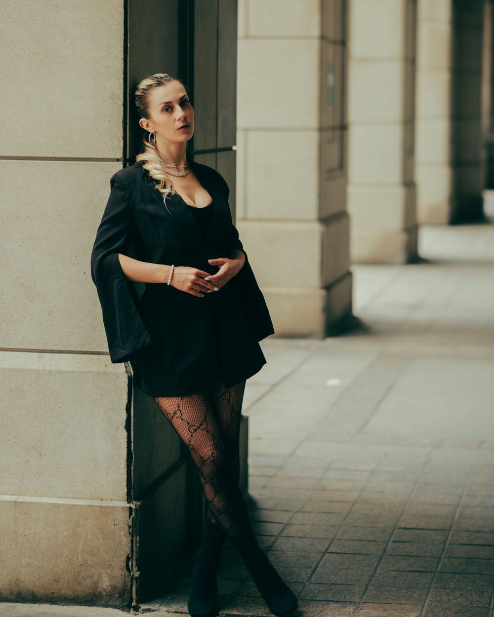 A person in a black dress and sheer stockings stands elegantly against a beige building column on an urban sidewalk.