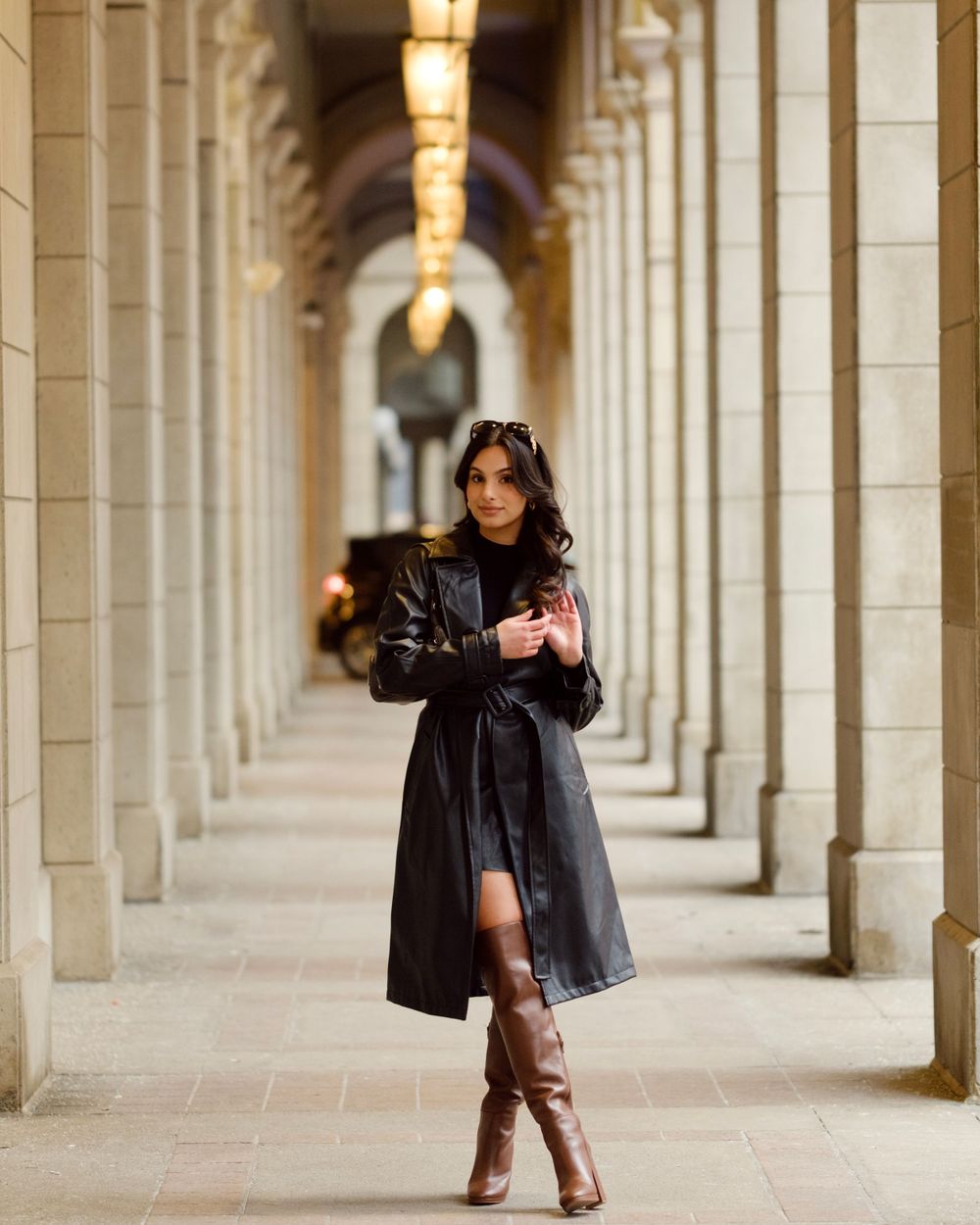 A woman in a black coat and boots poses in an elegant arched hallway with warm lighting.