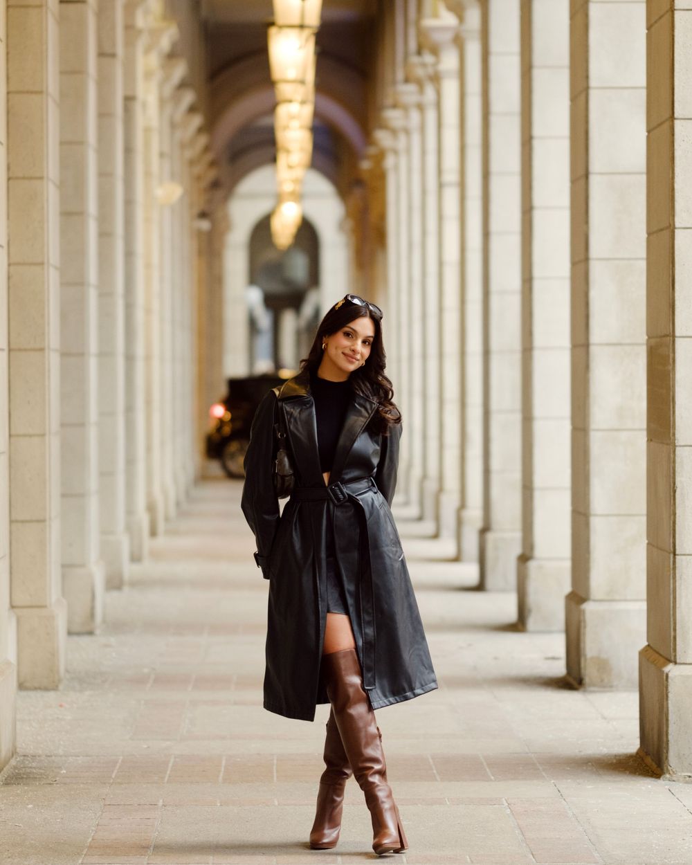 A person in a black coat poses in a long corridor with arched columns and warm lighting creating an elegant atmosphere.