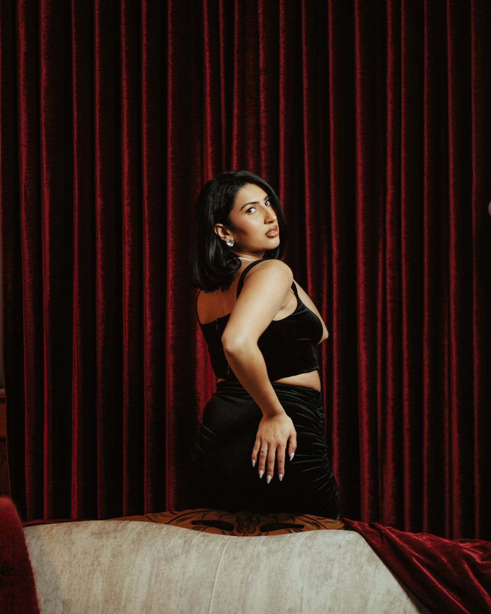 A woman in a black outfit poses elegantly against red velvet curtains in a dramatic theatrical setting.