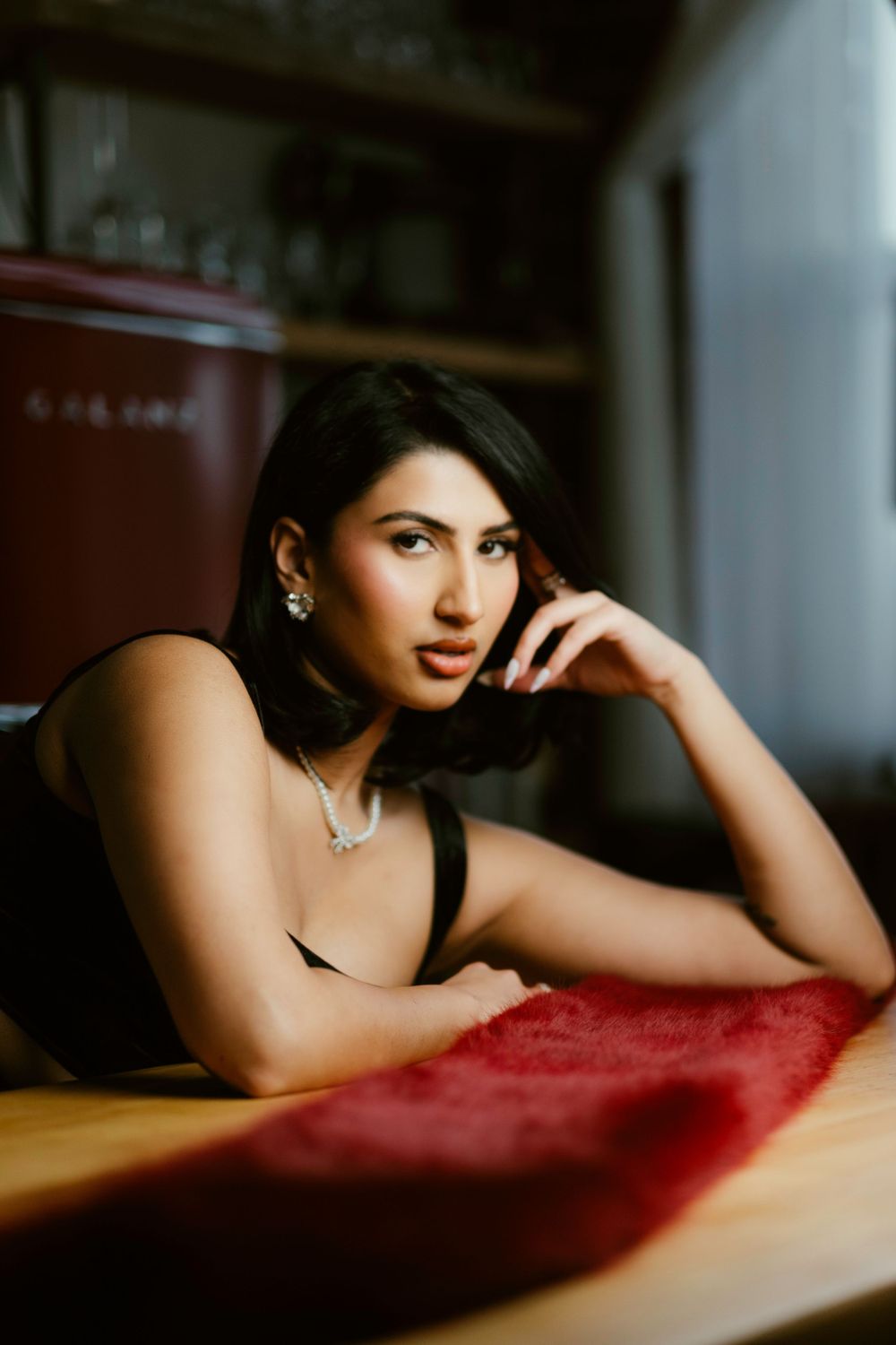Woman in black evening wear with jewelry poses elegantly against red surface in moody lighting.