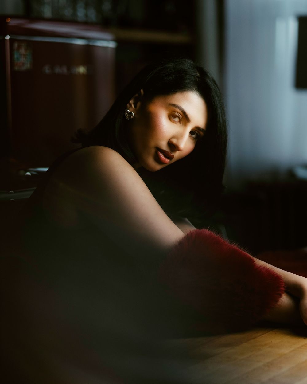 Moody portrait in dark lighting with dramatic shadows and red accents on figure leaning beside a wooden table.