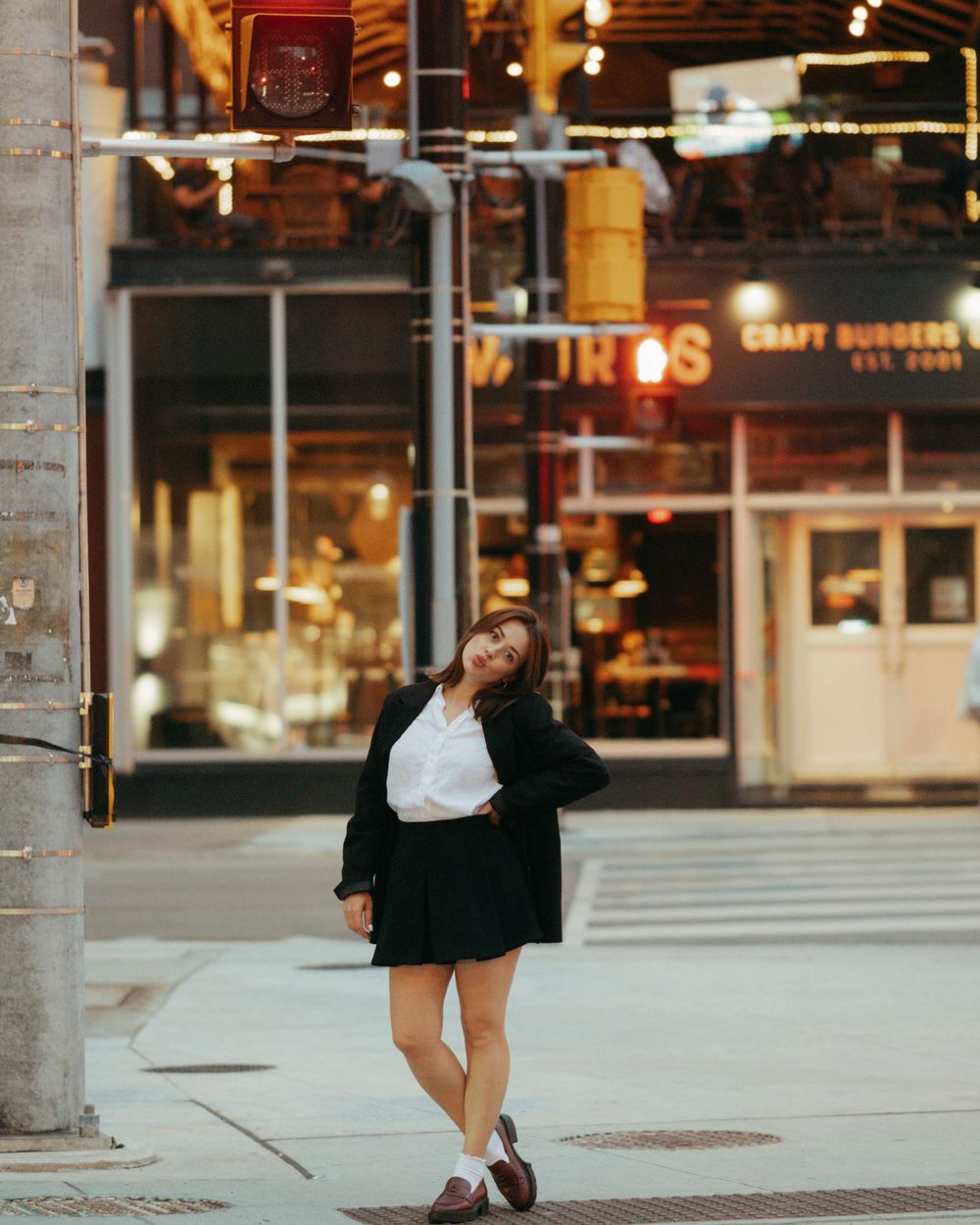 A person in a black jacket and white top poses playfully on a city street at dusk with storefronts in the background.