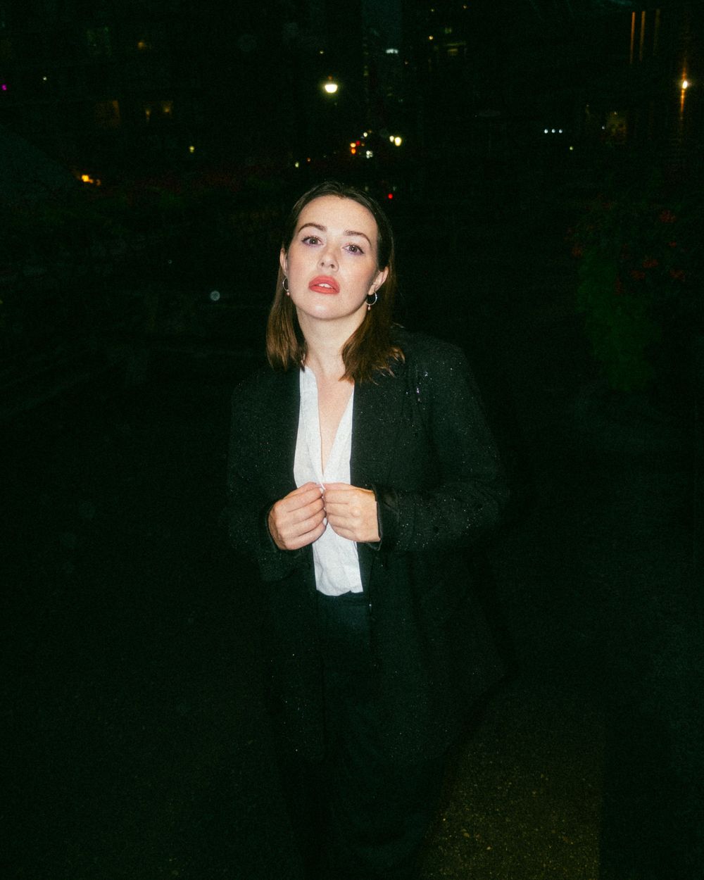 Stylish figure in a black blazer jacket and white shirt poses at night with city lights in the background.