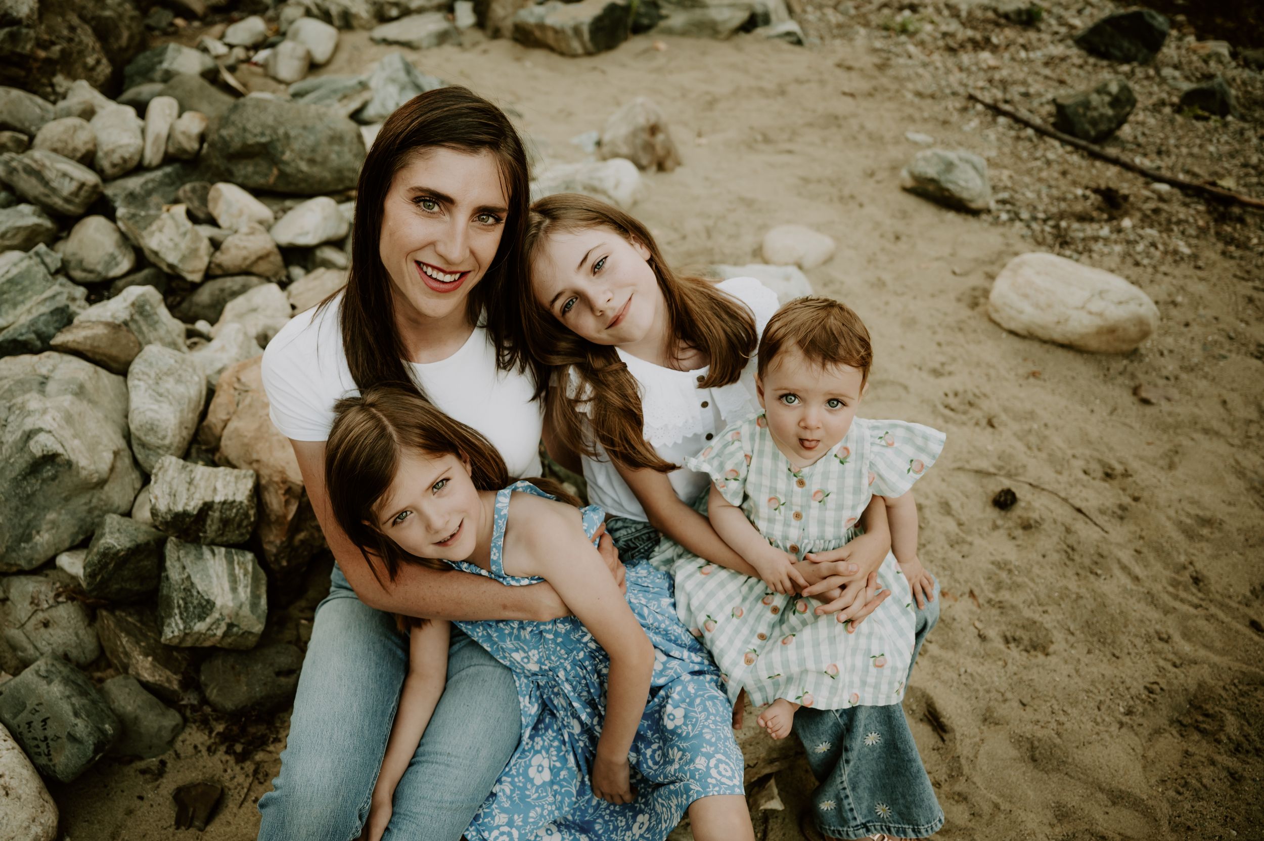 A mother smiles with her three young daughters while sitting on a sandy beach surrounded by rocks.