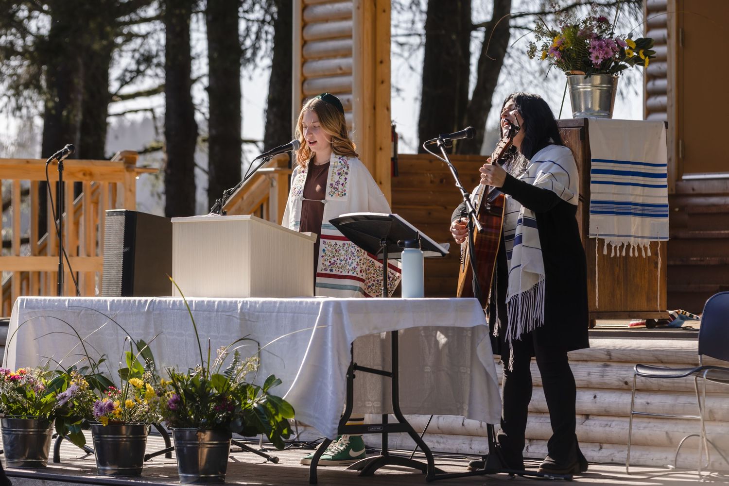 An outdoor Bat Mitzvah ceremony takes place on a rustic wooden deck with floral arrangements on the altar table.