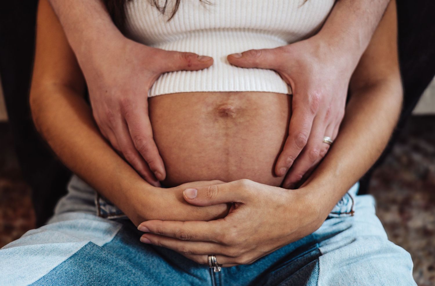 Two sets of hands gently hold a pregnant belly wearing a white sweater and blue jeans.