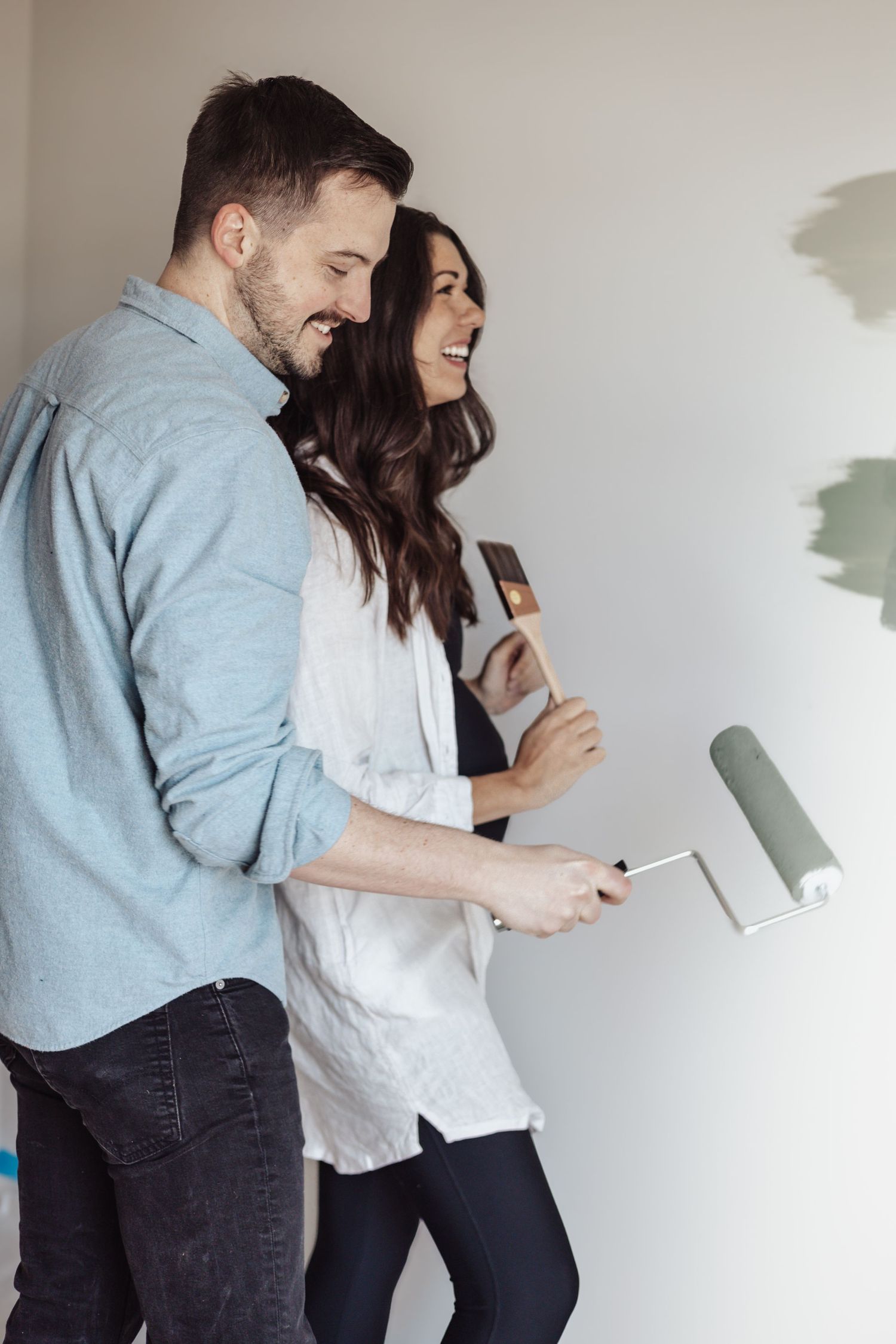 A couple laughs together while holding paint rollers during a home improvement project.