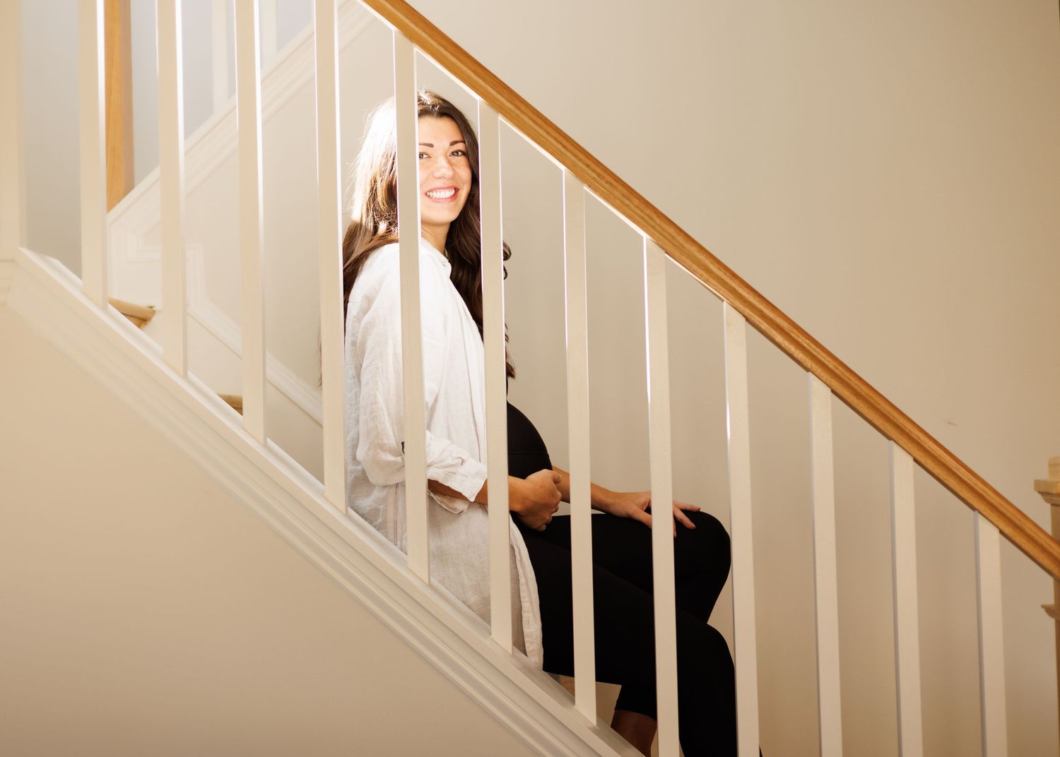 Person in white sweater sitting on carpeted stairs with wooden handrail and white balusters in a home.