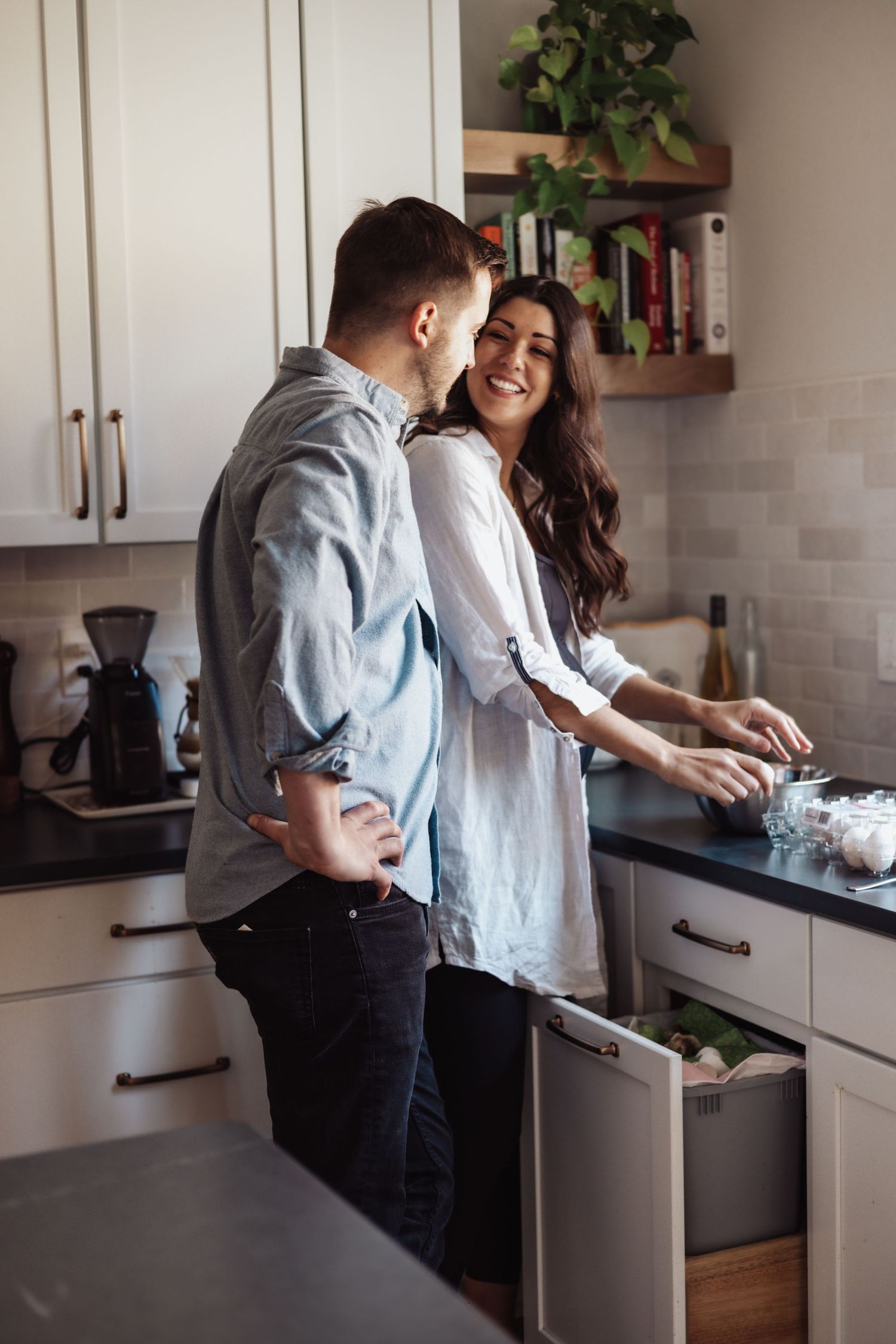 A couple shares a sweet moment while preparing food together in their modern white kitchen with houseplants on floating shelves.