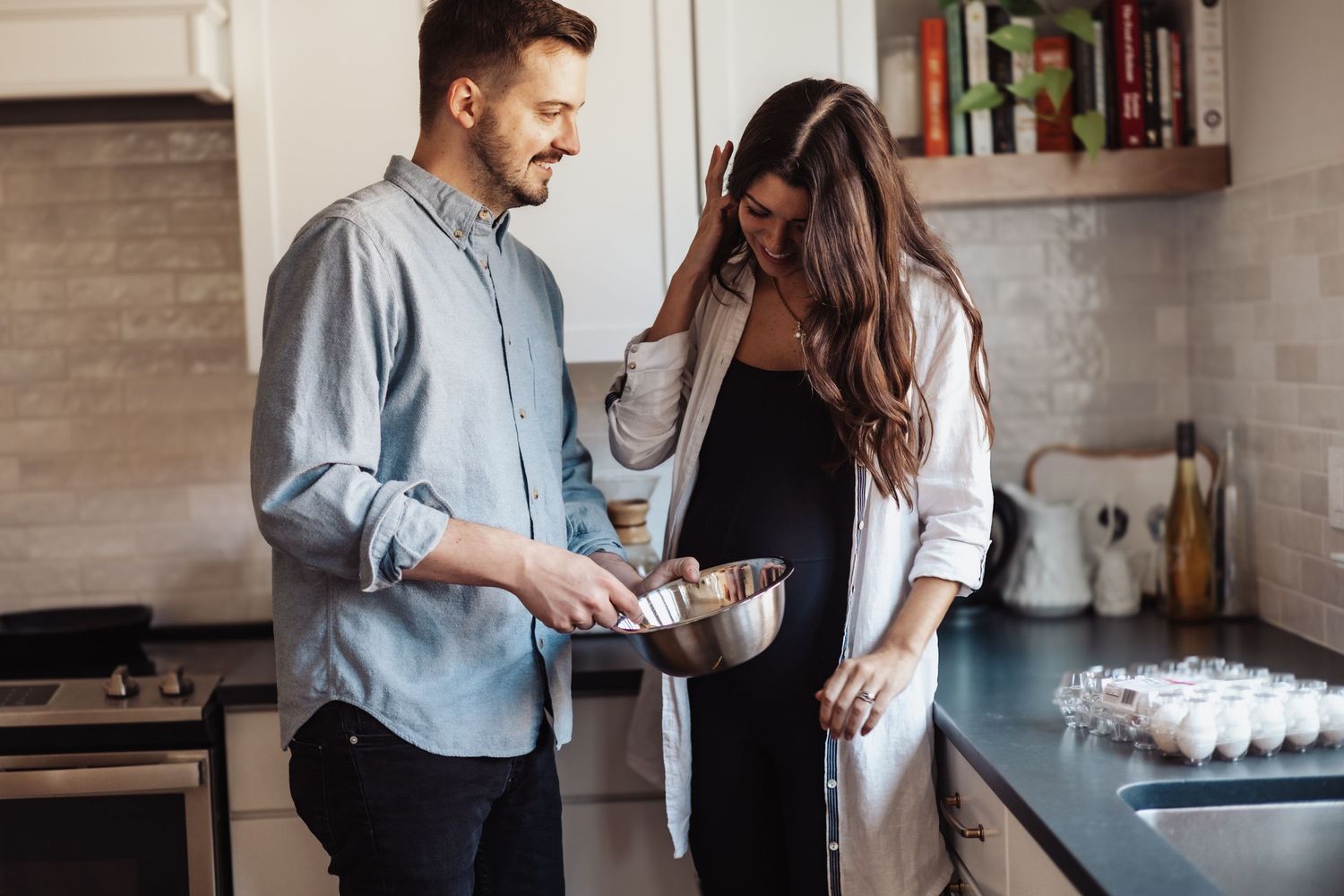 A couple wearing casual clothing laughs together while making coffee in their modern kitchen.