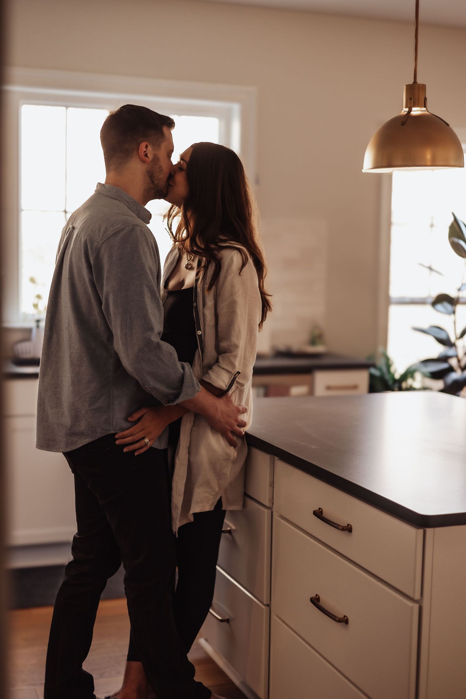 Romantic couple sharing an intimate moment in a modern white kitchen with pendant lighting and dark countertops.