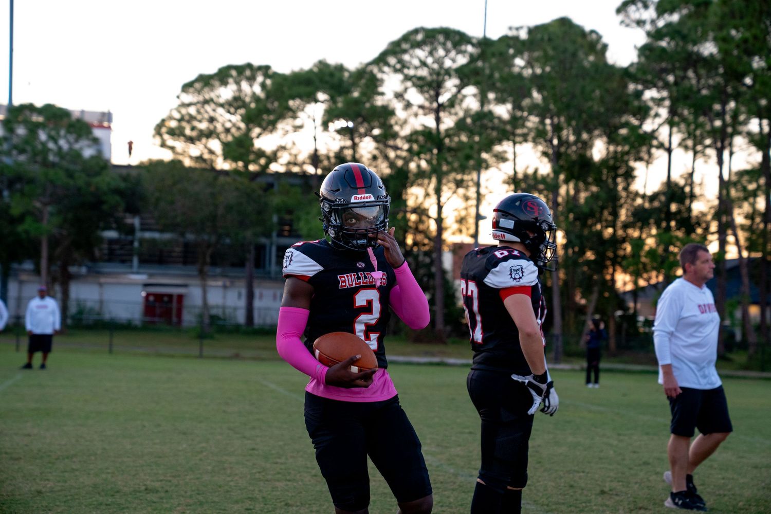 Football players in dark uniforms with pink accents practicing on field at dusk.
