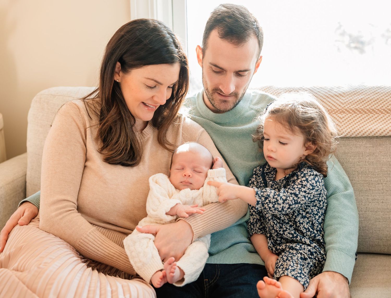 Parents sitting on couch with newborn baby and toddler in casual home setting.