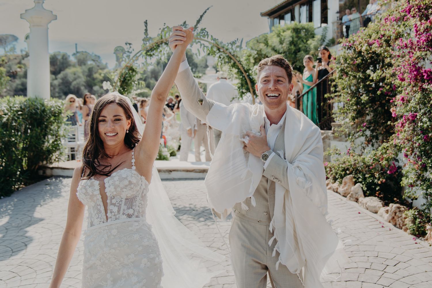 A joyful newlywed couple celebrates together on a sunny pathway with blooming pink flowers at an outdoor venue.