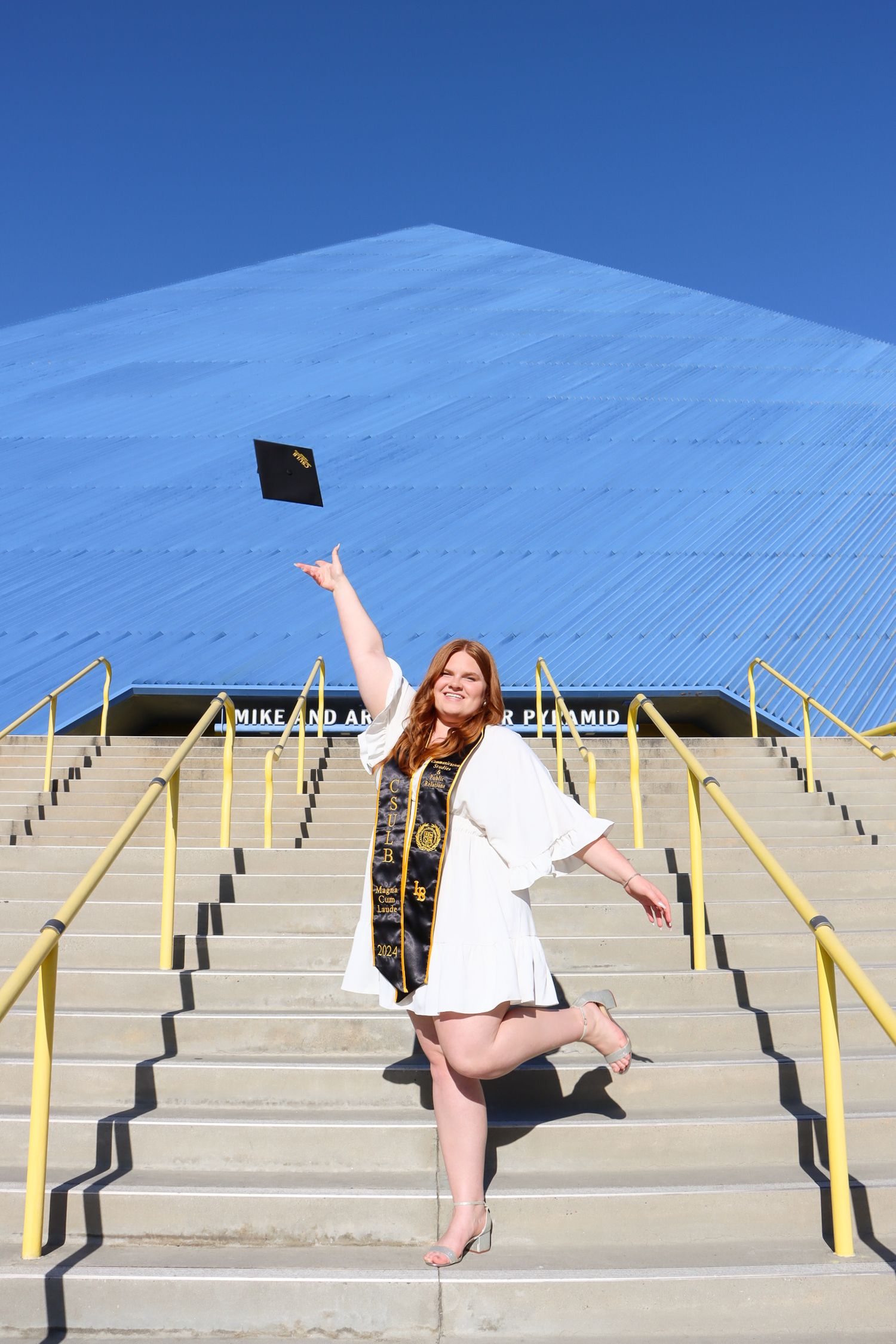 Graduate in white dress and academic regalia throws cap on concrete steps with blue wall background.