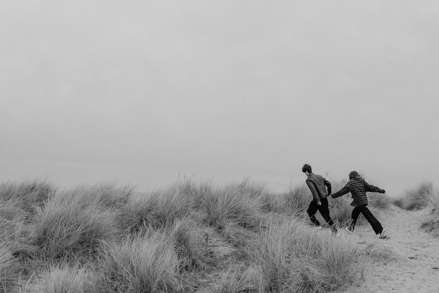 a black and white photo of a man leading a lady by her hand up some sand dunes at the beach.