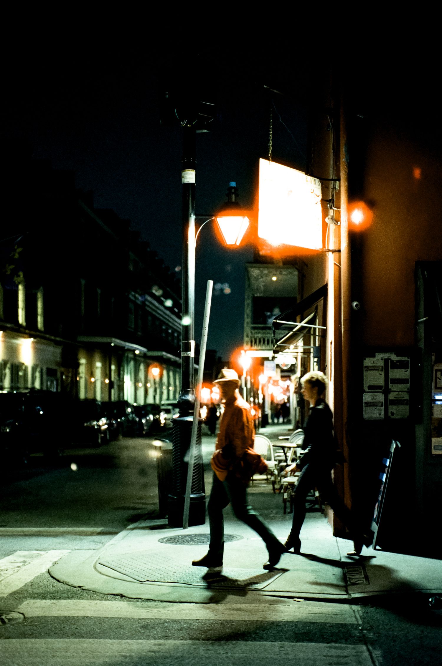 People walk along a dimly lit city street at night with glowing street lamps and storefronts creating an atmospheric scene.