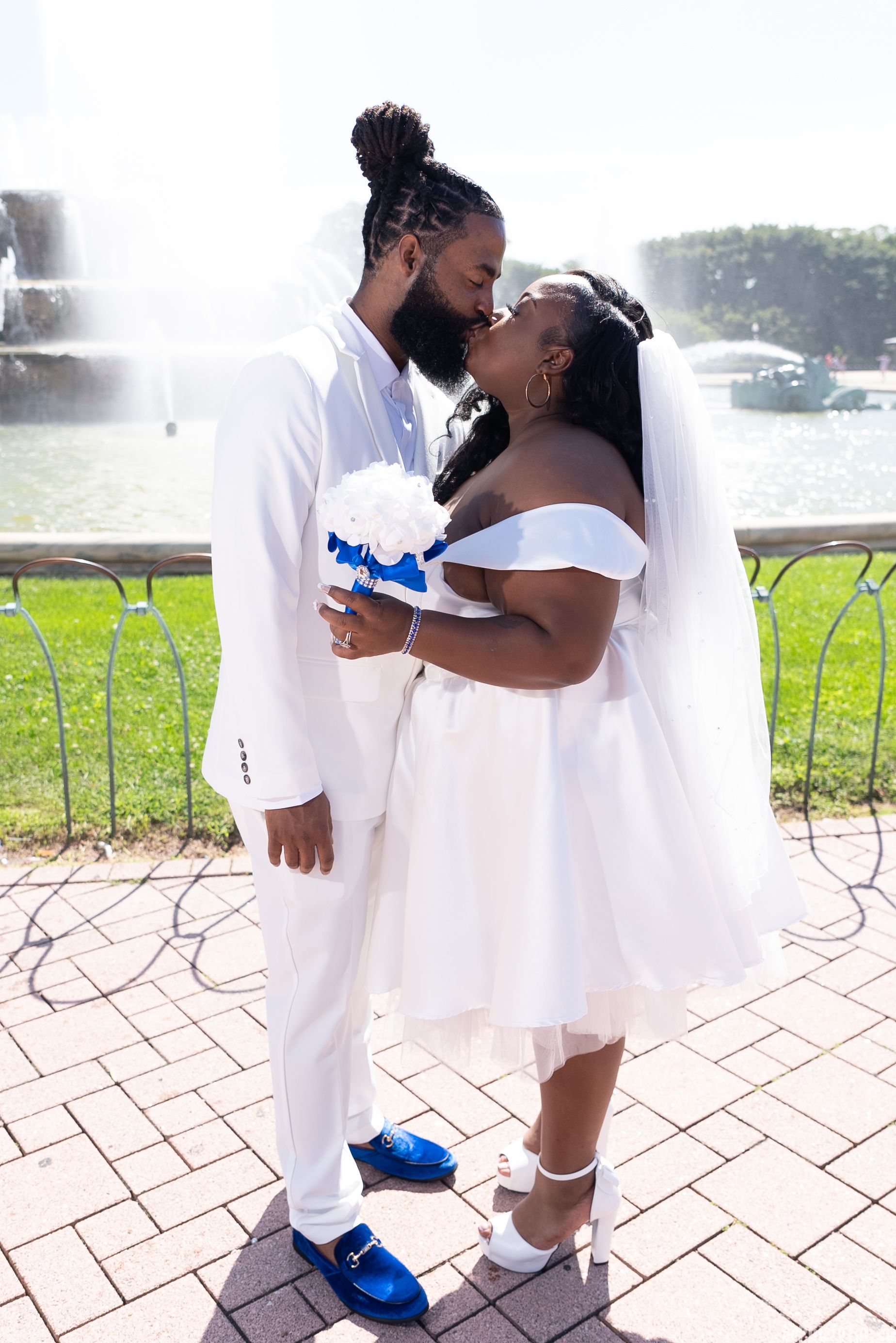 Newlywed couple in white outfits embracing near a fountain on their wedding day.