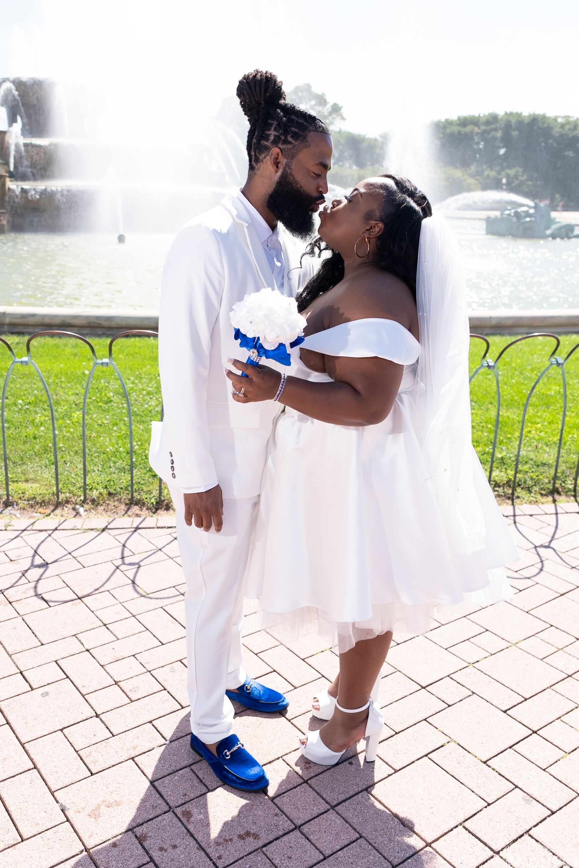 Wedding couple sharing intimate moment in white attire with bright blue shoes against fountain backdrop.