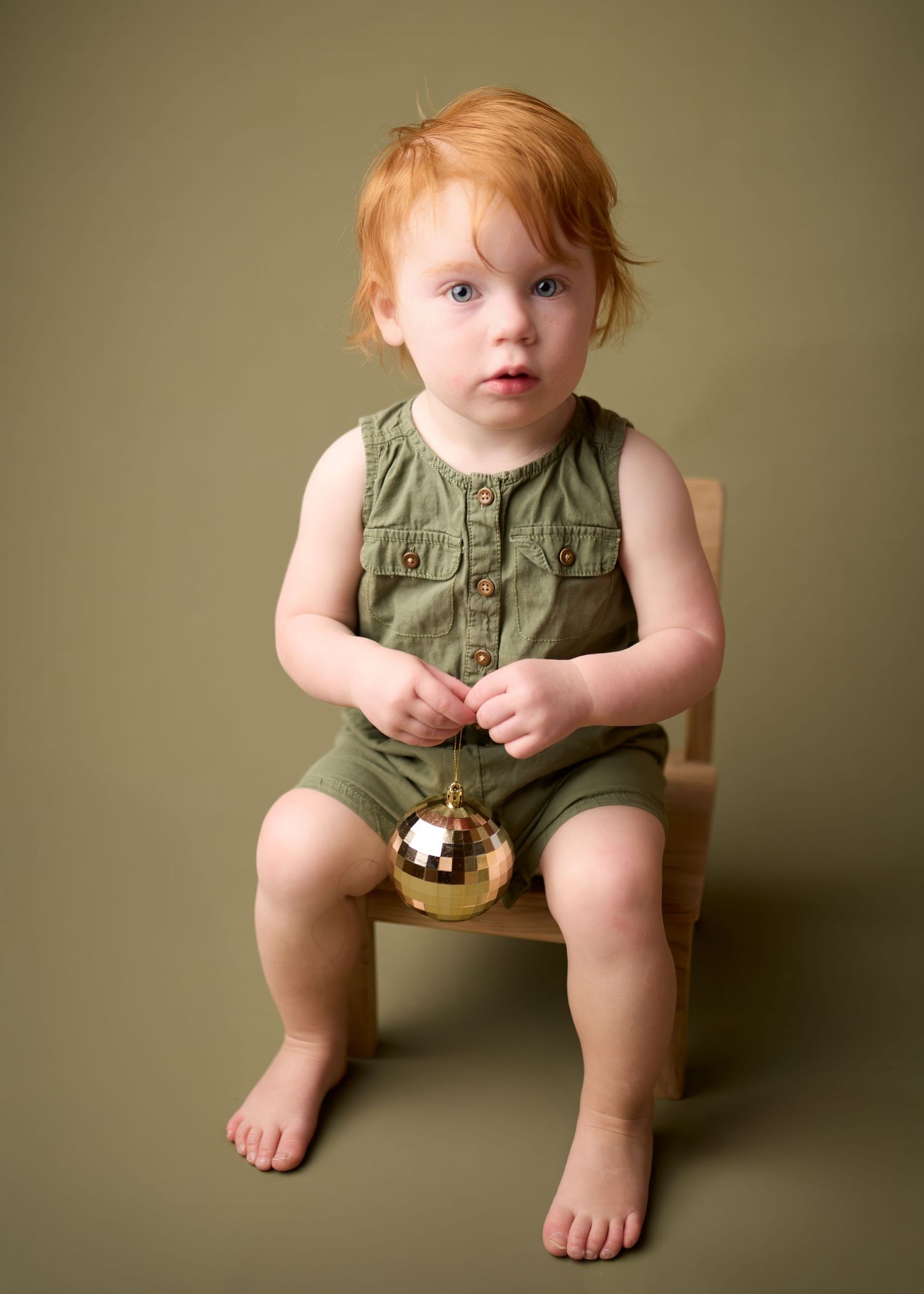 Small child in olive green overalls sits on wooden chair holding gold Christmas ornament against neutral backdrop.