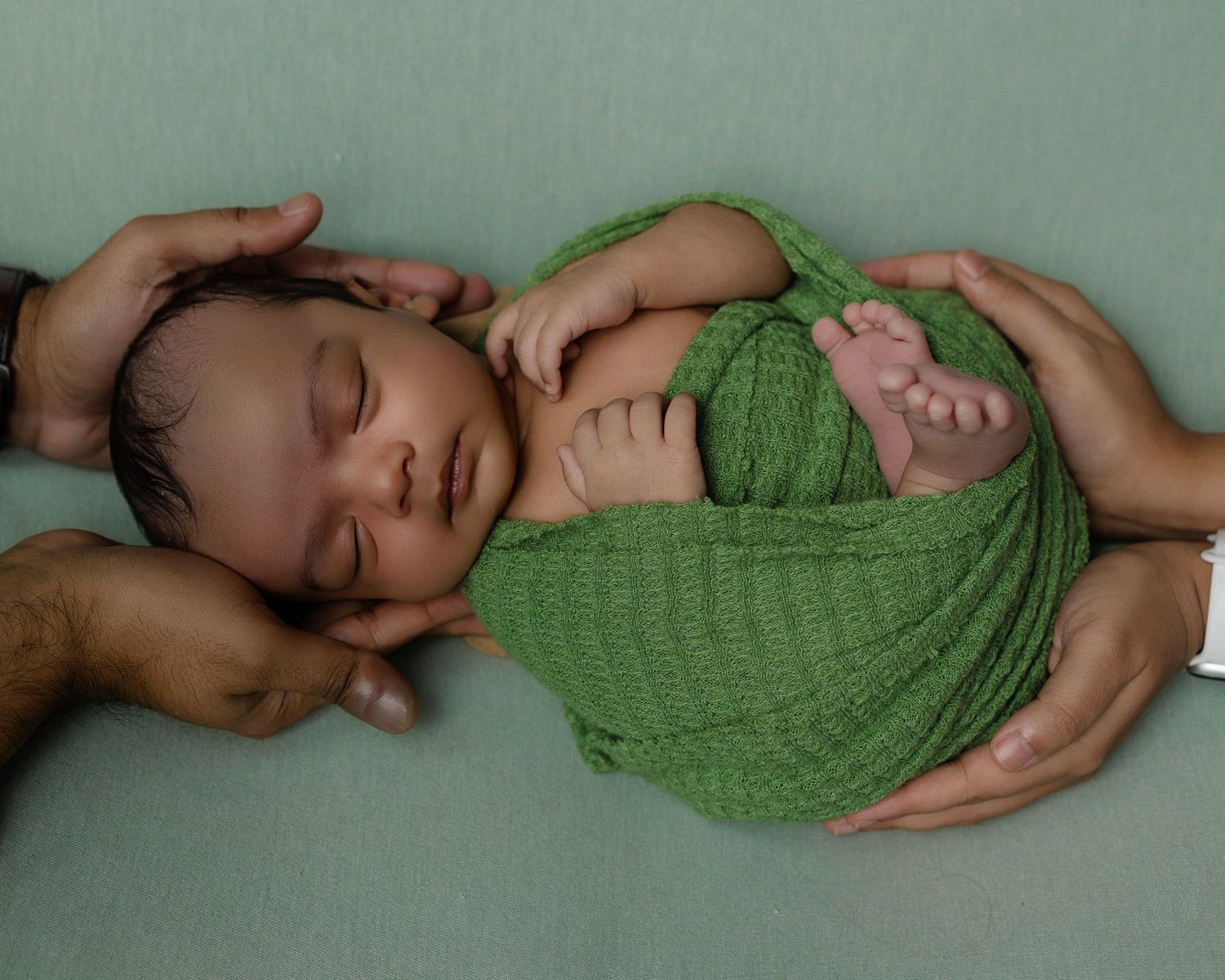 Newborn baby peacefully sleeping while wrapped in soft green knit blanket against sage background.