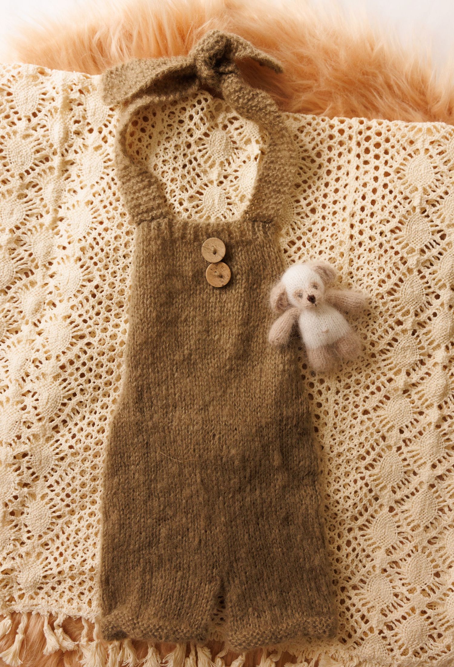 Handknit brown baby romper with lace trim and small teddy bear detail laid on cream-colored lace fabric.