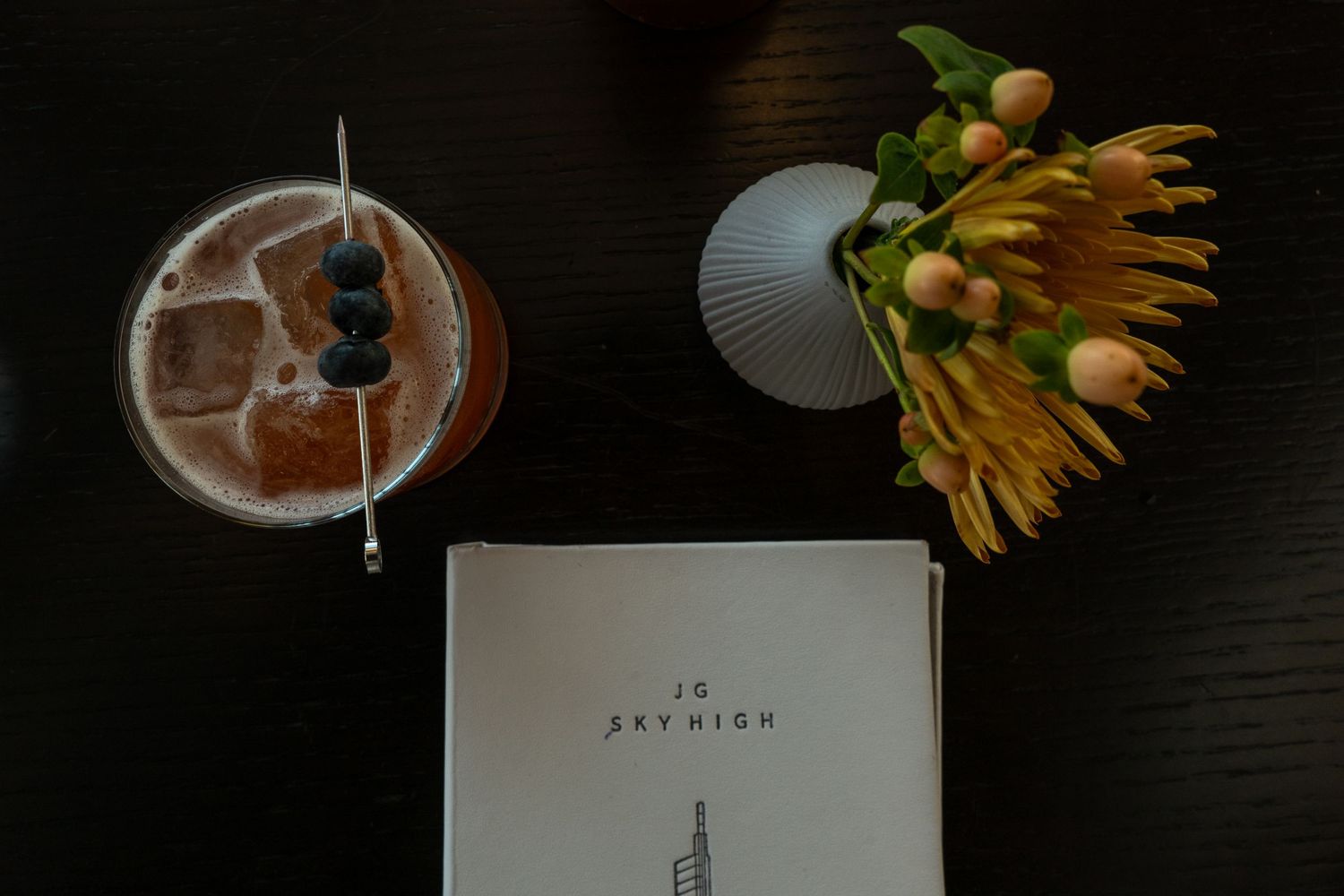 Flatlay with cold brew coffee, yellow sunflower bloom, and minimalist book on dark wooden surface.
