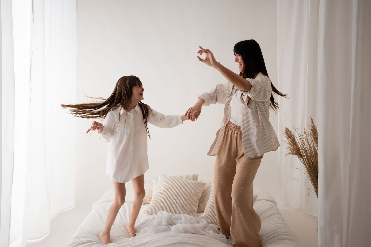 A mother and daughter dance and hold hands together on a white bed in a bright room.