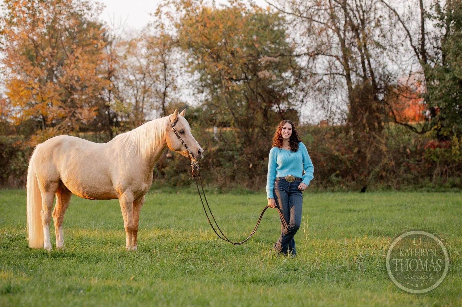 Someone in blue stands with a cream-colored horse in an autumn field.