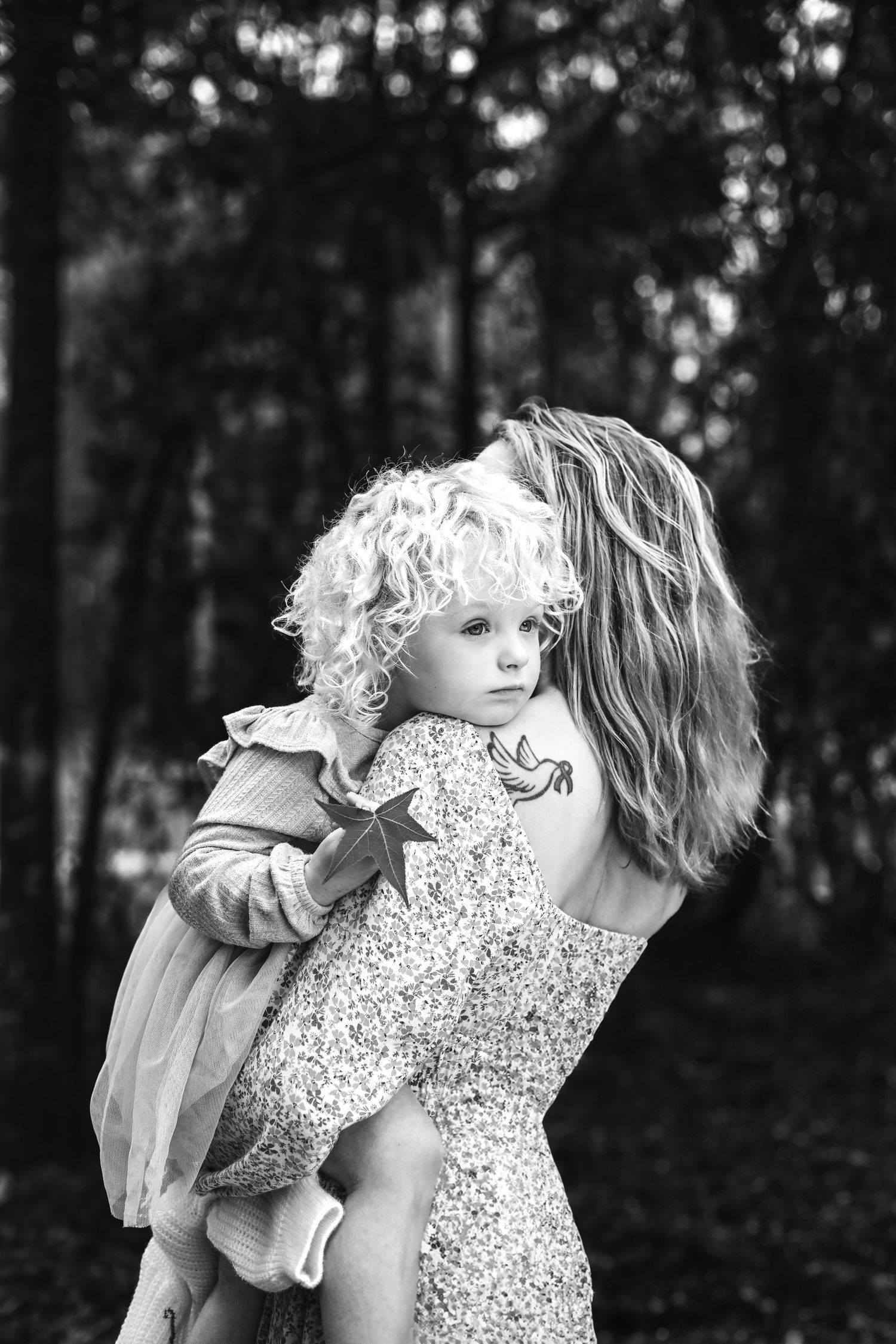A tender black and white portrait showing a small child with curly blonde hair being held in floral dress outdoors.