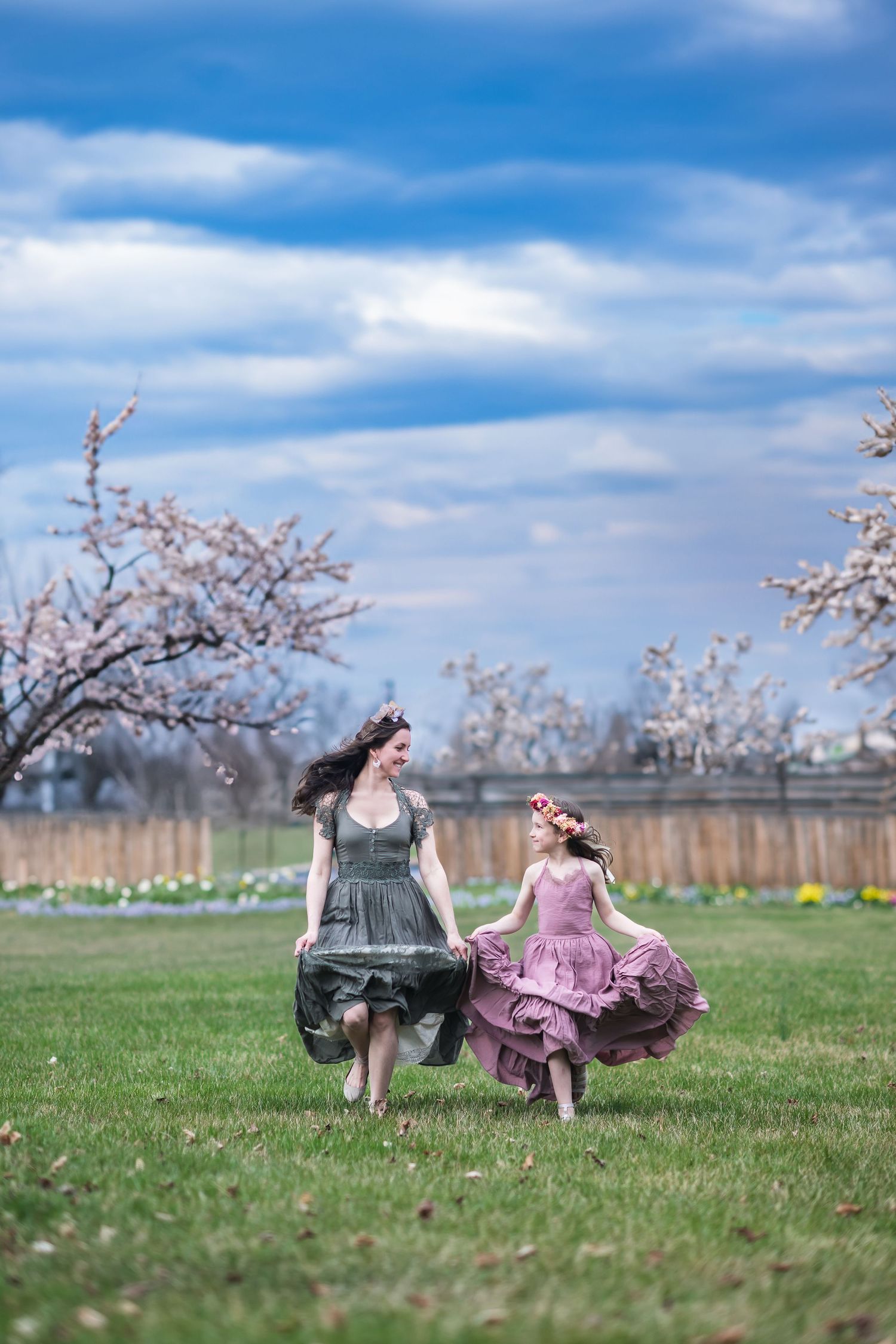 Two figures in flowing dresses twirl and dance together in a spring field with blooming trees in the background.