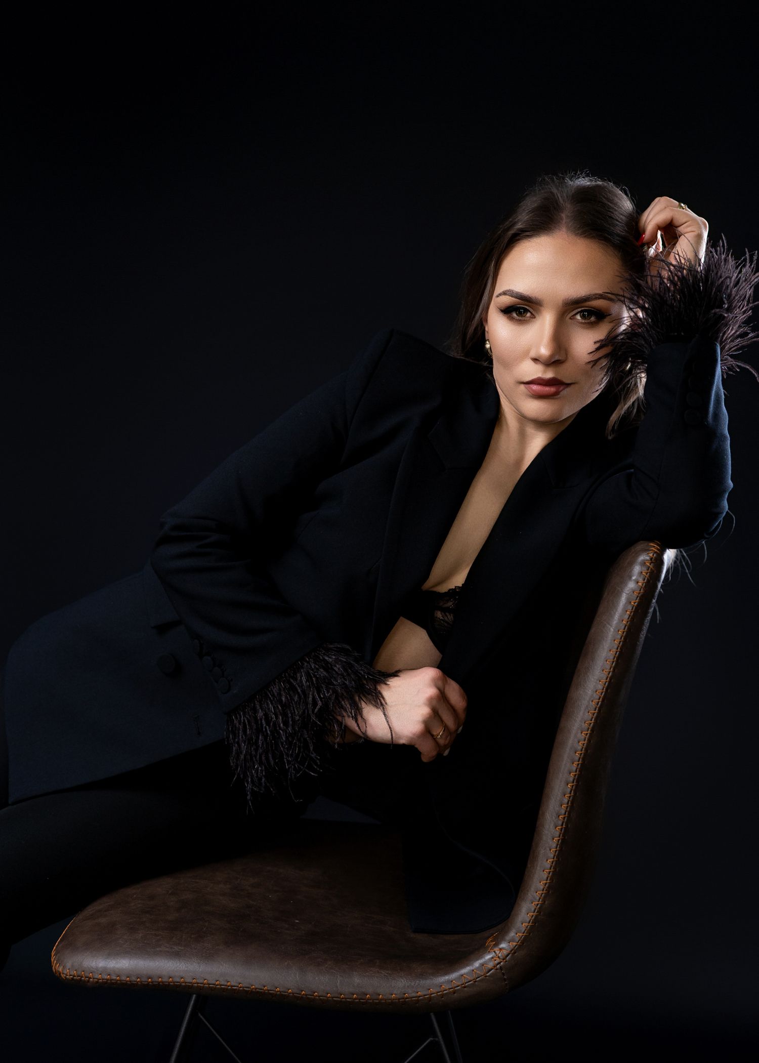 Model in black blazer reclines dramatically on brown leather chair against dark background.