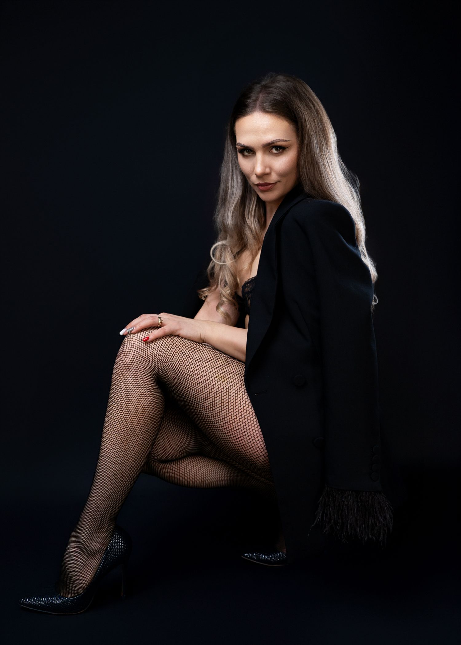Fashion model in black coat and fishnet stockings poses against dark backdrop.