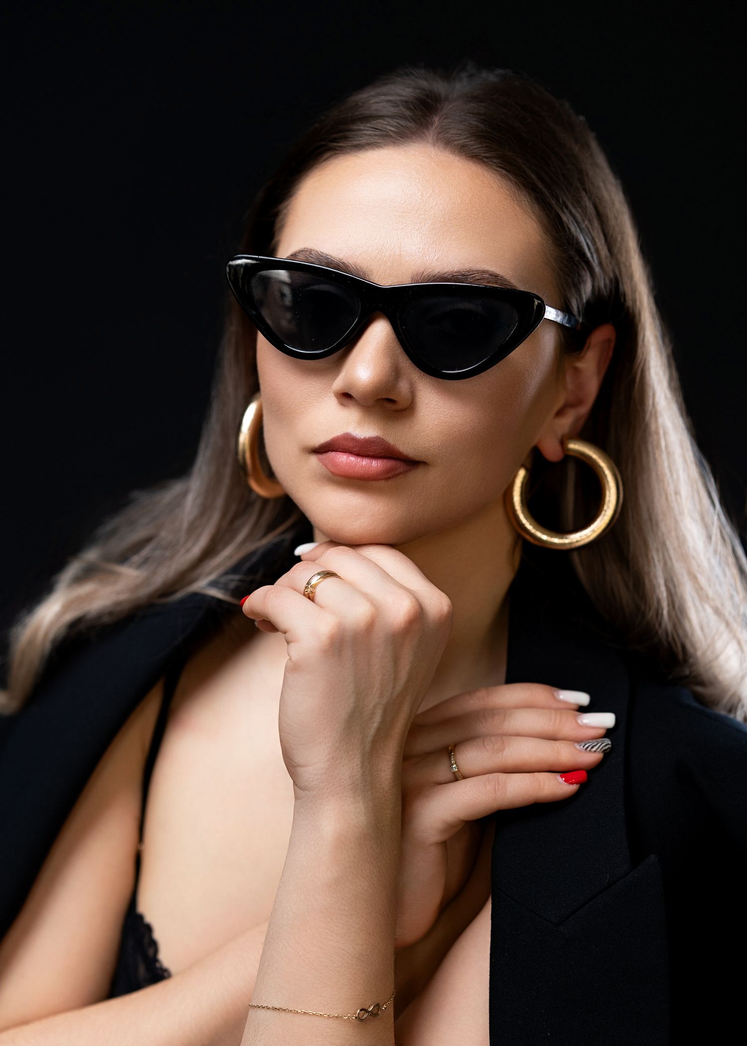 Stylish portrait with black sunglasses, gold hoop earrings and red nails against dark background.