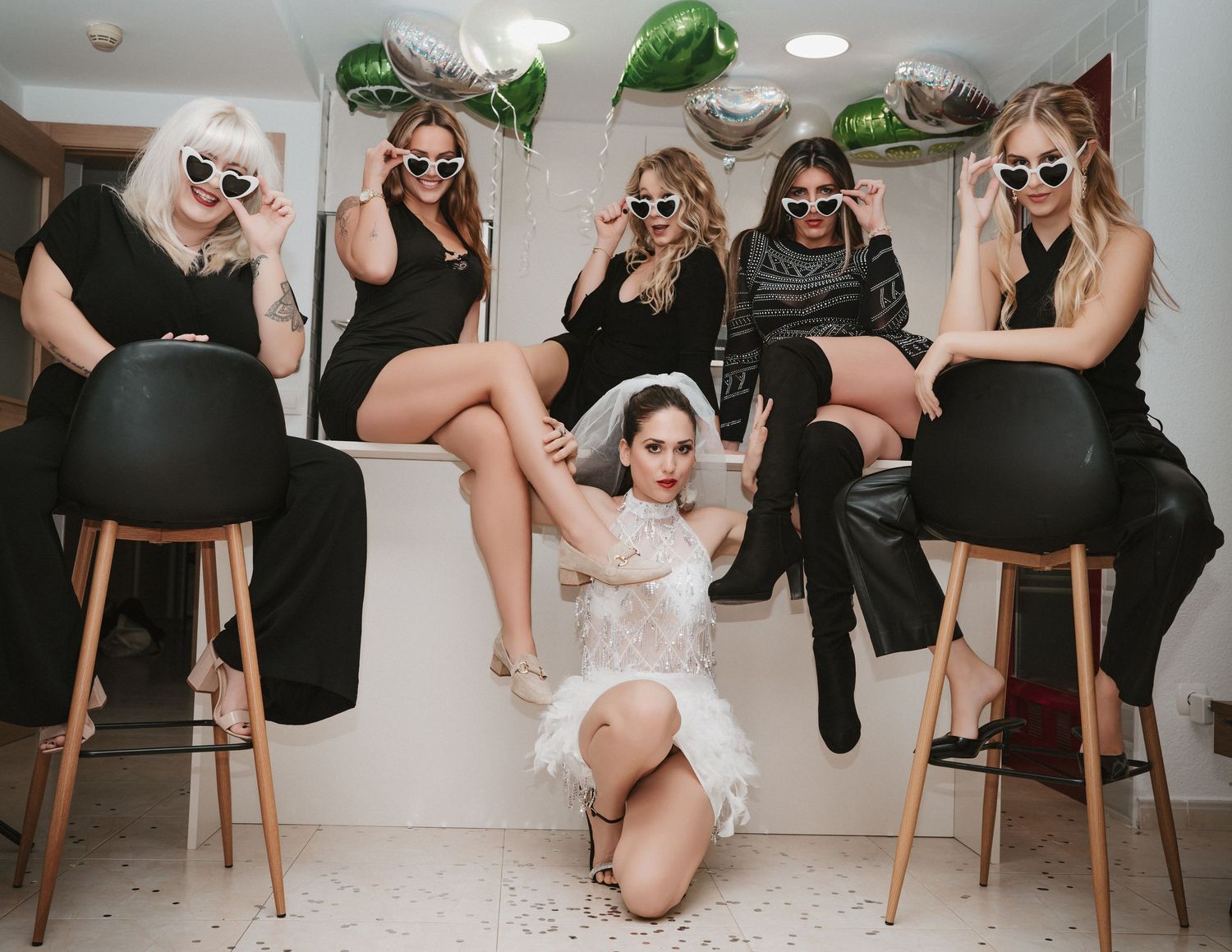 A group of friends in black outfits and sunglasses pose together with one person in white at a stylish indoor gathering.