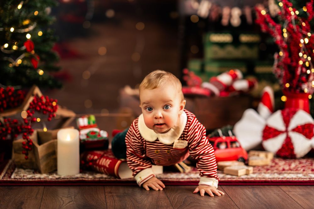 A baby in red and white stripes crawls on a wooden surface near Christmas decorations and festive holiday decor.
