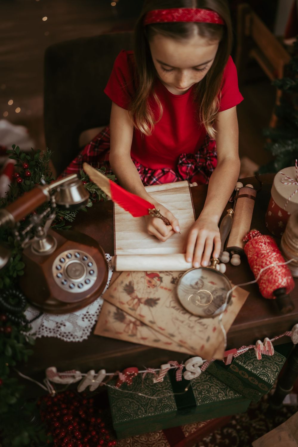 A person in a red dress writes a letter at a vintage desk with a rotary phone and holiday decorations nearby.