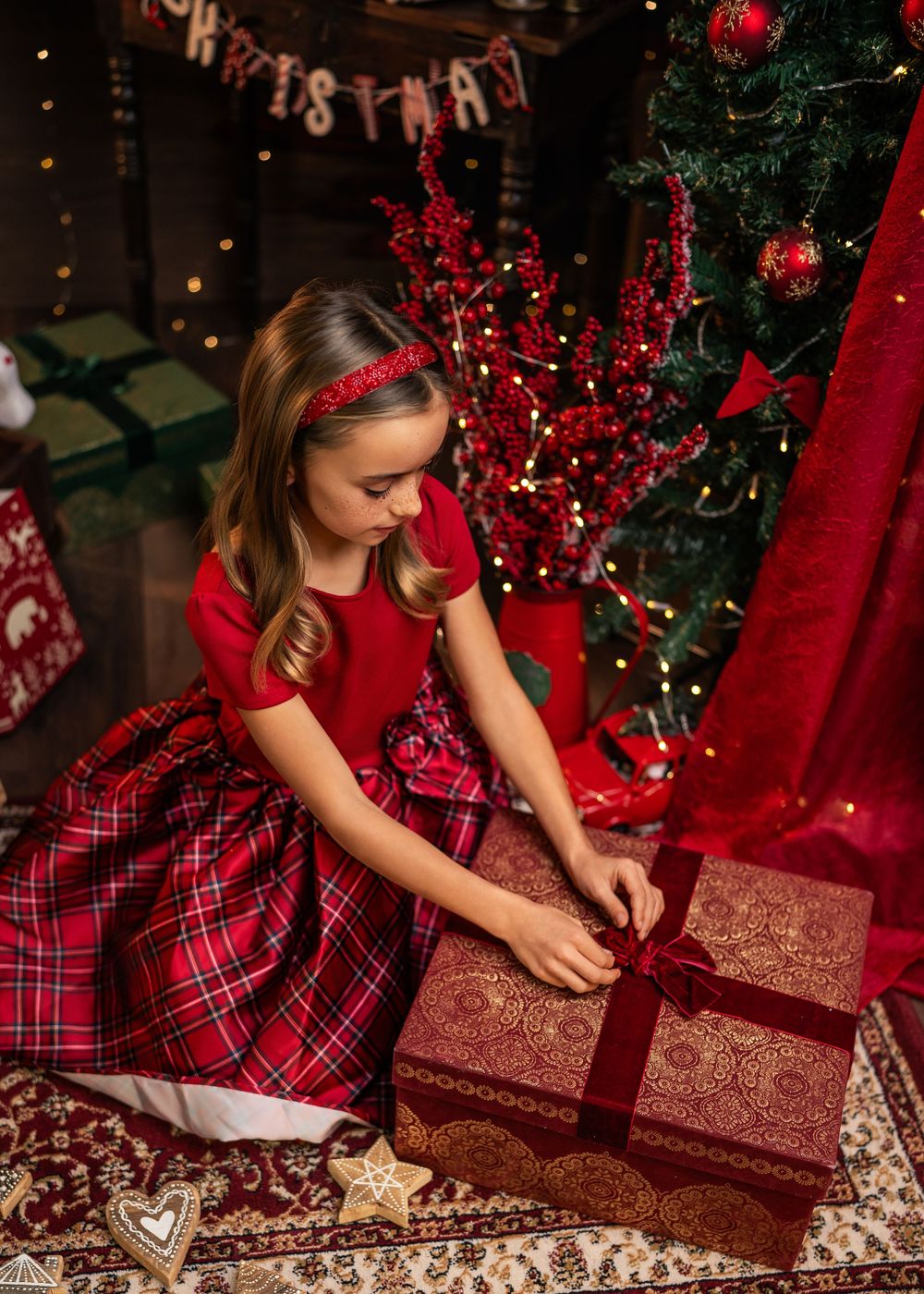 A person in a red plaid dress unwraps a sparkly red Christmas present near a decorated tree.