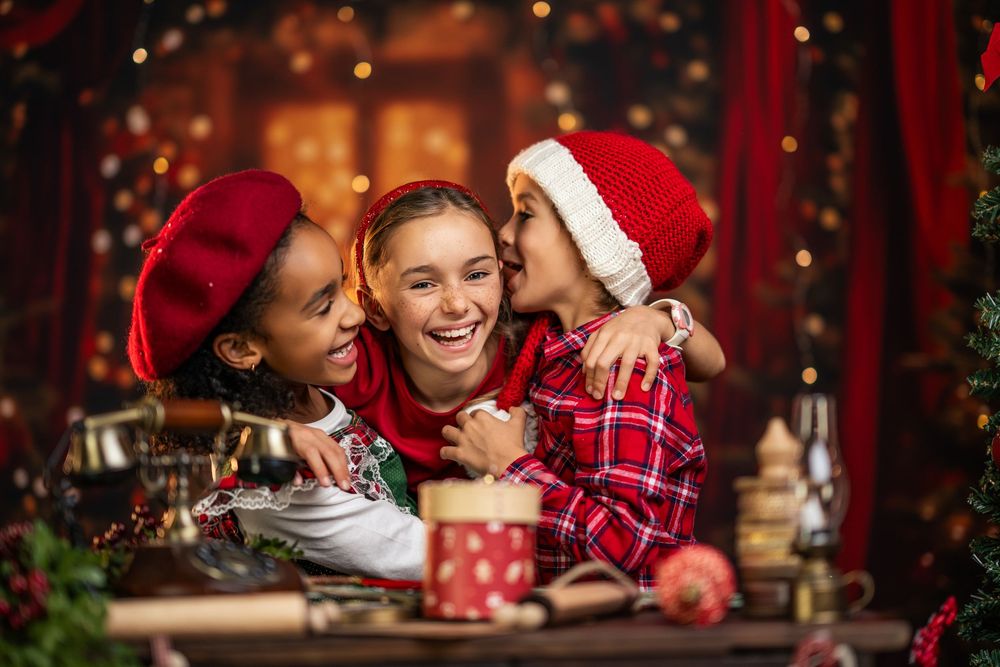 Three friends in festive holiday attire share joyful moments around a decorated Christmas table with hot drinks and treats.