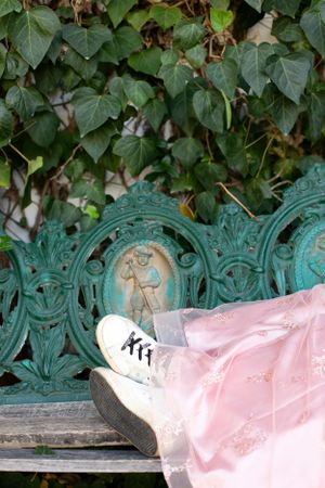 Pink dress fabric and white sneaker against an ornate green metal bench with ivy backdrop.