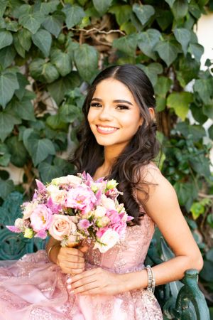 Person in a pink gown holding a bouquet of pink and peach flowers against leafy green backdrop.
