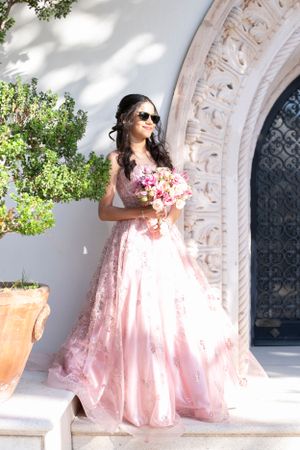 Someone in a flowing pink gown holding a bouquet against an ornate archway in bright sunlight.